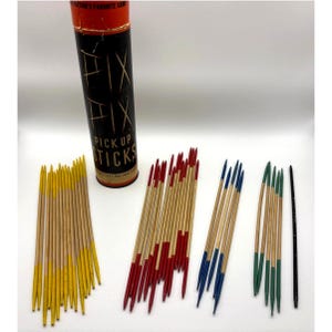 Può includere: Un set di gioco mikado vintage composto da un tubo cilindrico in cartone con etichette nere e arancioni e la scritta FIX FIX PICKUP STICKS. Il set include numerosi bastoncini di legno con punte dipinte in giallo, rosso, blu e verde.