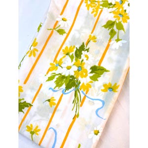 May include: White fabric with a pattern of yellow and white daisies, green leaves, and blue ribbons. The fabric has vertical orange stripes. The daisies are clustered in bouquets and scattered across the surface.