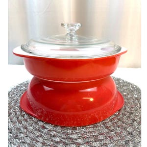 Puede incluir: Una fuente para horno roja con tapa de cristal transparente y borde blanco. El plato está colocado sobre una base roja a juego. La tapa tiene un pomo de cristal transparente. El plato es probablemente vintage y adecuado para cocinar y servir.