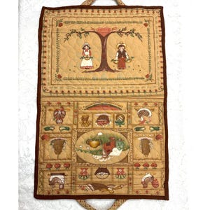 May include: A quilted wall hanging with a brown border and a beige background. The top section features a tree with two children holding baskets. The bottom section has a grid of various images, including a house, a rooster, and a basket of fruit.