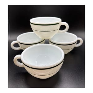 May include: Four white ceramic coffee cups with a black stripe design. Each cup has a rounded handle and a black stripe around the rim. The cups are stacked on top of each other against a black background.