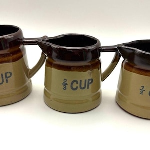 May include: Three ceramic measuring cups with handles and spouts, featuring a two-tone brown and tan glaze. The cups are labeled with black text reading 1 CUP, 2/3 CUP, and 1/2 CUP, indicating their respective liquid capacities.