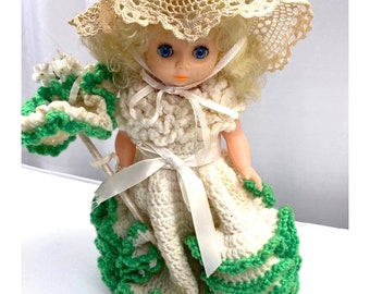 Op de afbeelding: Een vintage pop die een witte gehaakte jurk draagt met groene versiering en een bijpassende gehaakte hoed. De pop heeft blond haar en blauwe ogen.