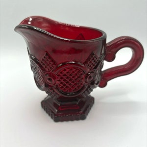 Puede incluir: Una jarra de crema de vidrio roja con un patrón de diamante y un asa. La jarra tiene una base de pedestal.