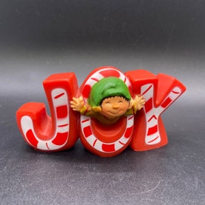 May include: Red, white and green Christmas JOY sign with a candy cane design. A small elf figure is peeking out from the centre of the O.