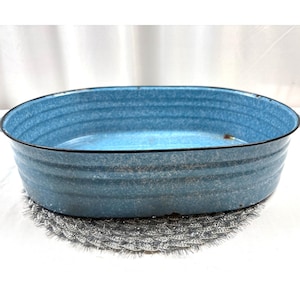 Könnte beinhalten: Ovale, hellblaue Emaille-Schüssel mit schwarzem Rand. Das Becken hat horizontale Rillen und steht auf einer silberfarbenen, strukturierten Matte. Die Emaille hat ein Vintage- und rustikales Aussehen.