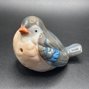 May include: A ceramic bird figurine with a light brown body, blue and gray wings, and a small orange beak. The bird has a small hole in its body.