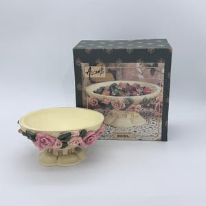 May include: A cream-colored pedestal bowl featuring a decorative band of sculpted pink roses and green leaves around the exterior. The bowl is displayed in front of its original dark green box, which features the text Accents and BOWL.