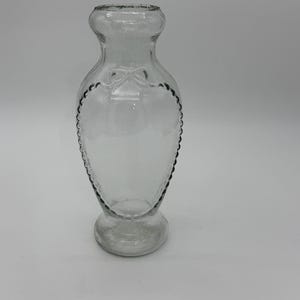 May include: Clear glass vase with a heart-shaped design. The vase has a flared base, a narrow neck, and a rounded top. A bow detail is visible on the front. The edges of the heart shape are outlined with a textured pattern.