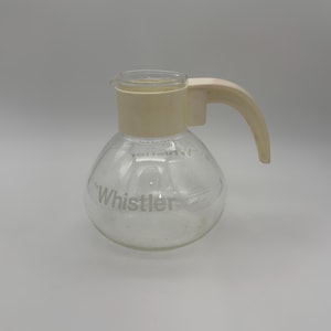Puede incluir: Una jarra de cristal transparente con cuerpo redondeado y un cuello y asa de plástico de color crema. El texto "The Whistler" está impreso en blanco en la parte frontal, con texto blanco adicional visible cerca del borde superior.