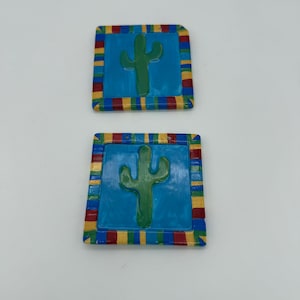 May include: Two square ceramic ornaments with a Southwestern theme. Each ornament features a green cactus on a blue background, framed by a colorful border of red, yellow, and blue stripes. The ornaments have small holes for hanging.