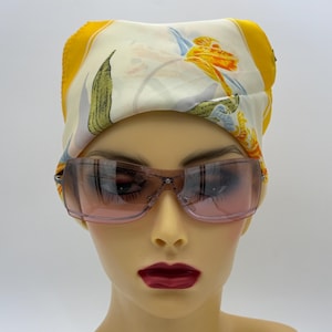 May include: A mannequin head wearing a cream-colored scarf with a yellow border and floral print, paired with pink-tinted sunglasses. The sunglasses have a clear frame and a dark pink lens.