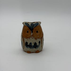 May include: A ceramic owl figurine with a tan body, brown eyes, and blue accents. The owl has a small opening at the top and a whimsical design. The figurine is a decorative item.