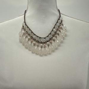 May include: A layered necklace with a bronze chain and white, teardrop-shaped beads. The necklace is displayed on a white mannequin, showcasing its cascading design. The beads have a faceted cut, reflecting light.
