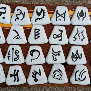 Pu&ograve; includere: Un set di 36 pietre triangolari bianche con simboli neri dipinti su di esse. Le pietre sono disposte in quattro file di nove pietre ciascuna. Le pietre sono su una superficie di legno.