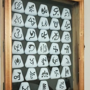 Pu&ograve; includere: Una scatola di legno con una parte anteriore in vetro mostra una collezione di 27 piastrelle triangolari bianche con simboli neri su di esse.