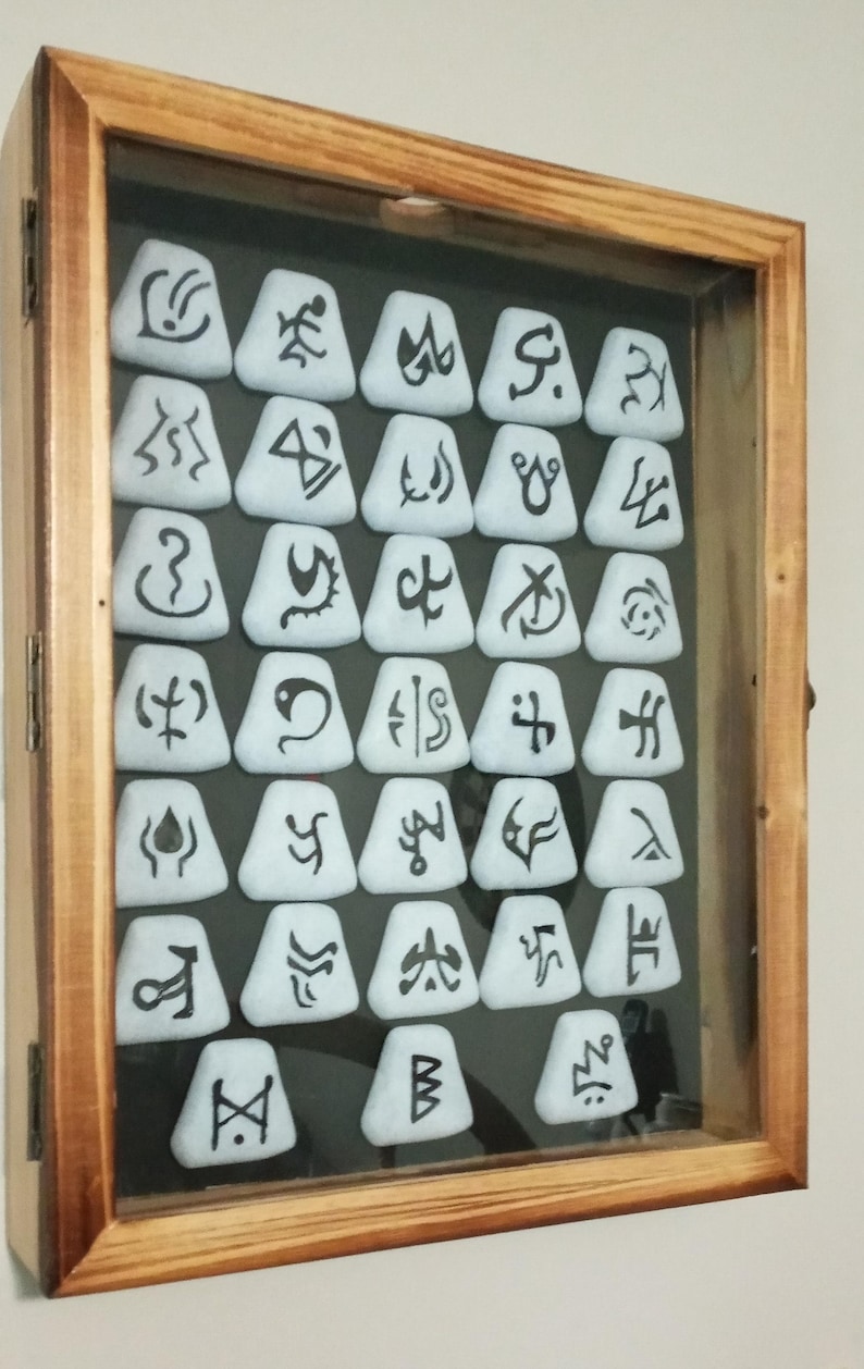 Pu&ograve; includere: Una scatola di legno con una parte anteriore in vetro mostra una collezione di 27 piastrelle triangolari bianche con simboli neri su di esse.