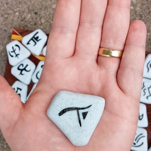 Pu&ograve; includere: Una mano tiene un sasso di forma romboidale, di colore azzurro chiaro e macchiato, con un simbolo nero su di esso. Il simbolo &egrave; una lettera "T" stilizzata con un piccolo triangolo in basso. Il sasso &egrave; circondato da altri sassi con diversi simboli su di essi.