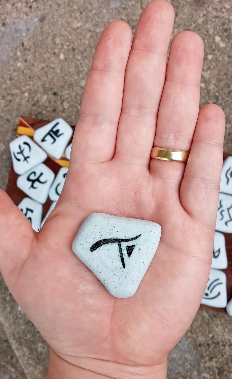 Pu&ograve; includere: Una mano tiene un sasso di forma romboidale, di colore azzurro chiaro e macchiato, con un simbolo nero su di esso. Il simbolo &egrave; una lettera "T" stilizzata con un piccolo triangolo in basso. Il sasso &egrave; circondato da altri sassi con diversi simboli su di essi.