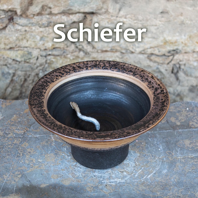 May include: A ceramic bowl with a dark interior and a decorative rim. The rim features a textured pattern in shades of brown and tan. The word "Schiefer" is displayed in white text above the bowl. A small, curved, silver-colored object rests inside.
