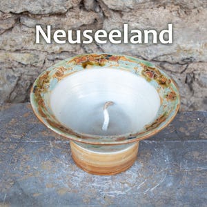 May include: A handmade ceramic candle holder with the text "Neuseeland". The candle holder has a wide, shallow bowl shape with a light green and brown rim. The base is a cylindrical shape with a light brown color.