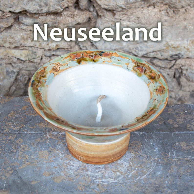 May include: A handmade ceramic candle holder with the text "Neuseeland". The candle holder has a wide, shallow bowl shape with a light green and brown rim. The base is a cylindrical shape with a light brown color.