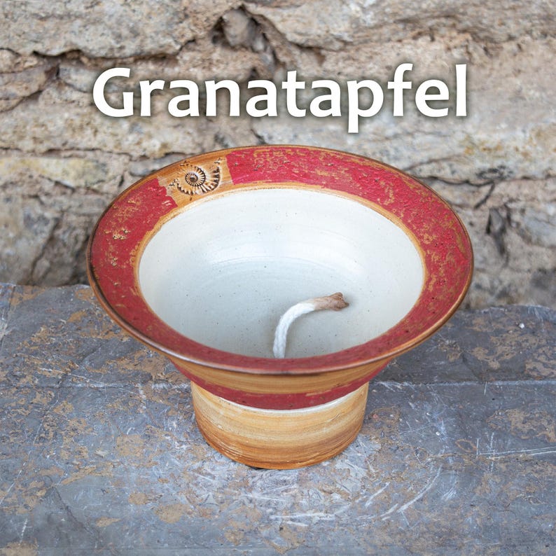 May include: A ceramic bowl with a red and gold rim and a white interior. The bowl has a wide, shallow design with a short, flared base. A white wick extends from the center. The word "Granatapfel" is printed on the top.