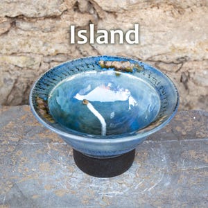 May include: A handcrafted ceramic bowl with a blue glaze, featuring a unique design and the word "Island" in white text. The bowl is supported by a dark base, set against a textured stone background. This decorative bowl is perfect for home decor.