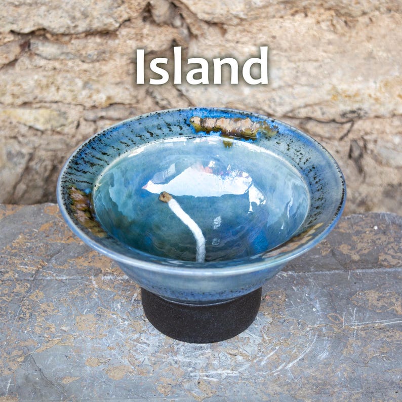 May include: A handcrafted ceramic bowl with a blue glaze, featuring a unique design and the word "Island" in white text. The bowl is supported by a dark base, set against a textured stone background. This decorative bowl is perfect for home decor.