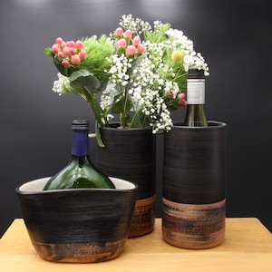 Könnte beinhalten: Drei schwarze Keramikvasen mit brauner Glasur am Boden. Die Vasen sind mit weißen und rosa Blumen gefüllt. Eine Vase hält eine Flasche Wein.