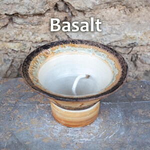 May include: A handmade ceramic candle holder with the word "Basalt" in white text. The candle holder has a white interior, a brown and tan rim, and a tan base. A white wick is visible inside the holder.