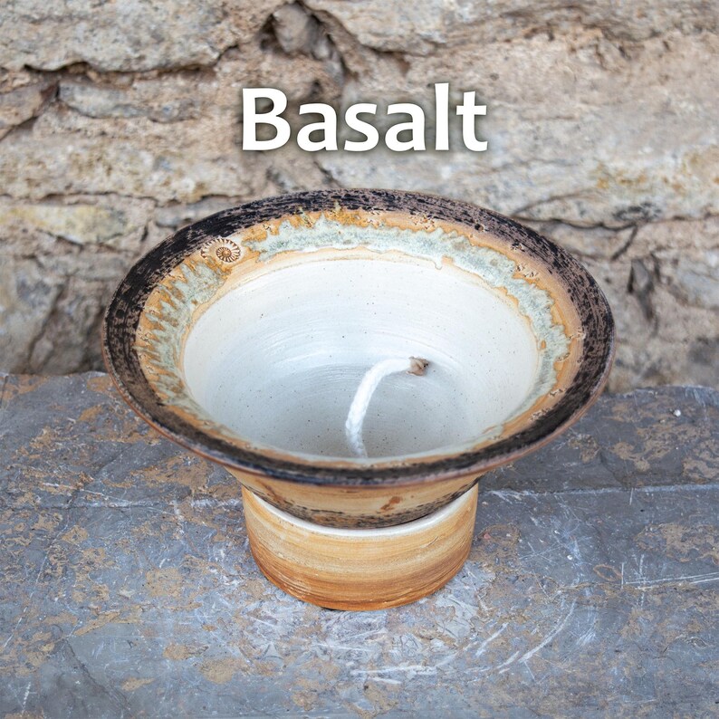 May include: A handmade ceramic candle holder with the word "Basalt" in white text. The candle holder has a white interior, a brown and tan rim, and a tan base. A white wick is visible inside the holder.