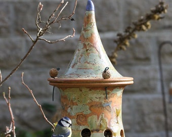 Op de afbeelding: Een blauwe mees zit op een keramische vogelvoederbak in de vorm van een toren met een puntdak. De voederbak heeft een groene, bruine en oranje glazuur met een blauwe bovenkant. De voederbak heeft meerdere gaten zodat vogels toegang hebben tot voedsel.