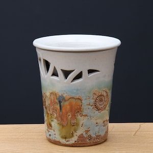 Könnte beinhalten: Weiße Keramikvase mit einem Ausschnittmuster um die Oberseite und einem blauen, grünen und orangefarbenen Glasurmuster. Die Vase hat eine strukturierte Oberfläche und zeigt ein Design aus versteinerten Muscheln.