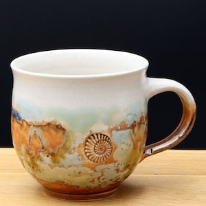 Könnte beinhalten: Eine weiße Keramiktasse mit braunem Henkel und einem einzigartigen Design mit brauner, orangefarbener und grüner Glasur mit einem erhabenen Ammoniten-Fossil-Design.