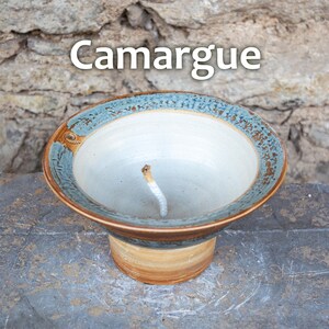 May include: A ceramic ashtray with the word "Camargue" in white text. The ashtray has a wide, shallow bowl with a blue and brown rim. The base is a light brown color, and a cigarette butt is in the center.