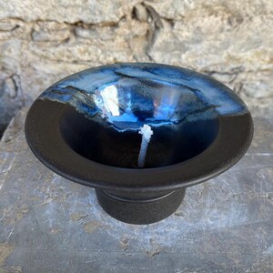 May include: A handmade ceramic candle holder with a deep blue and black glaze. The bowl-shaped holder has a wide rim and a small base. A white wick is centered in the bowl. The candle holder is on a stone surface.