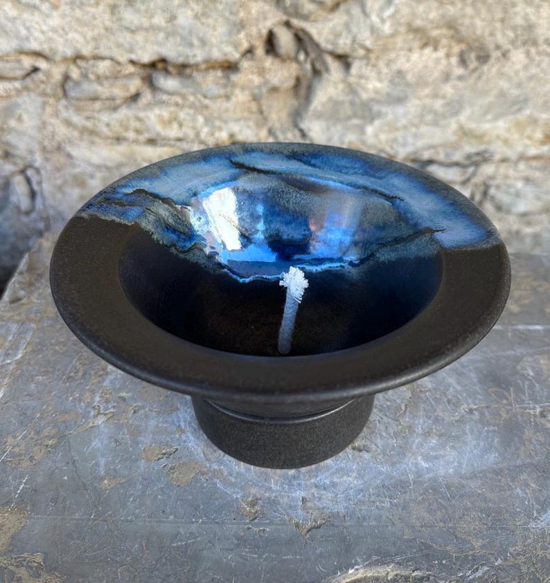 May include: A handmade ceramic candle holder with a deep blue and black glaze. The bowl-shaped holder has a wide rim and a small base. A white wick is centered in the bowl. The candle holder is on a stone surface.