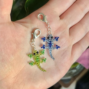 May include: Two beaded lizard charms. One is green and yellow, the other is silver and blue. Each charm has a silver clasp. The lizards are made of small beads and have detailed legs and eyes.