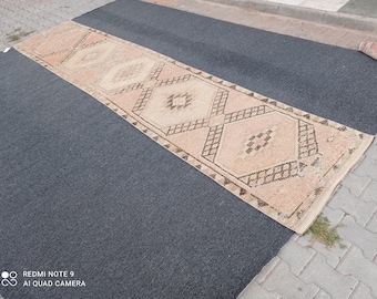 May include: A long, rectangular rug with a beige background and a geometric pattern in brown. The rug has a diamond shape with smaller diamonds inside.