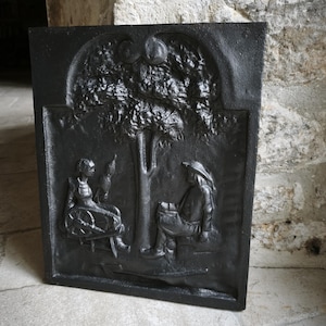 Peut inclure: Insert de cheminée en fonte noire avec une scène en relief de deux personnages sous un arbre. L'un est assis à un rouet, l'autre lit. L'arbre a deux gros fruits.