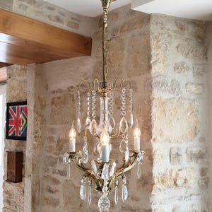 Op de afbeelding: Een messing kroonluchter met drie kaarsvormige gloeilampen, gebogen metalen armen en talloze heldere glazen kristallen druppelprisma's. Het armatuur hangt aan een decoratieve messing ketting en een plafondkap met een klassiek metalen frame.