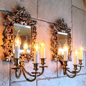 May include: Two ornate, gold-colored wall sconces with mirrors and candle holders. Each sconce features a rectangular mirror framed by intricate floral designs. White candles with flame-shaped bulbs are in the holders, creating a warm, inviting ambiance.