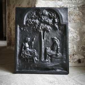 May include: A black cast iron fireplace insert featuring a detailed relief scene. The scene shows a woman with a spinning wheel and a man seated under a tree. The tree has fruit, and a bird is perched on the woman's hand.