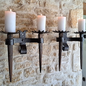 May include: Three wrought iron wall sconces with candle holders. Each sconce has a pointed, spear-like arm and a decorative star-shaped base. The sconces are mounted on a stone wall and each holds a white candle.