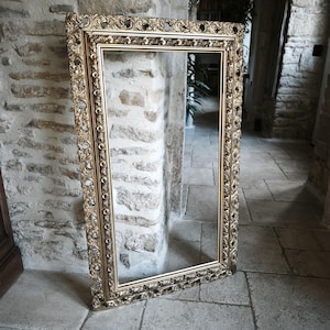 Può includere: Una grande cornice rettangolare dorata e ornata. La cornice presenta intricati dettagli floreali e a volute. Lo specchio è appoggiato a un muro di pietra in un corridoio con pavimento in pietra.