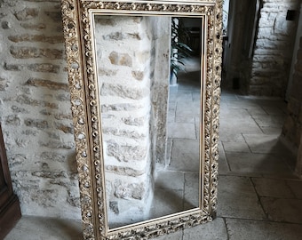 May include: A large, rectangular, ornate gold-colored mirror frame. The frame has intricate floral and scrollwork detailing. The mirror is leaning against a stone wall in a hallway with a stone floor.