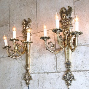 Peut inclure: Deux appliques murales dorées ornées, chacune avec trois lumières en forme de bougie. Les appliques présentent des motifs décoratifs floraux et aviaires. Elles sont fixées sur un mur de pierre clair texturé.