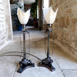 Könnte beinhalten: Zwei antike Tischlampen mit floralem Sockel und schlankem Stiel. Die Lampen haben eine Bronzefinish und sind mit einem weißen, tulpenförmigen Schirm versehen. Die Lampen sind über ein braunes Kabel an eine Stromquelle angeschlossen.