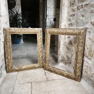 May include: Two ornate, gold-colored picture frames with detailed floral and scrollwork designs. The frames are rectangular and empty, leaning against a stone wall. The frames are approximately 24 inches tall and 20 inches wide.